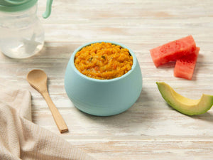 Bowl of stage 3 pork and apple roast dinner baby food with mashed puree, avocado slice, and watermelon pieces on a wooden surface.
