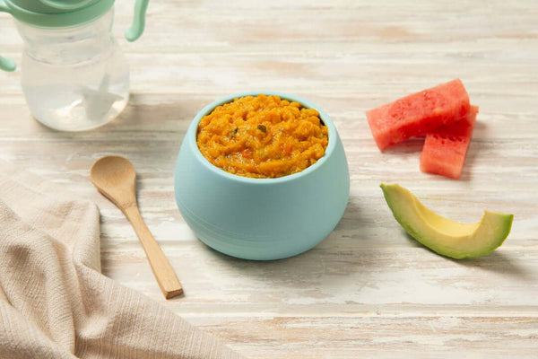 Bowl of stage 3 pork and apple roast dinner baby food with mashed puree, avocado slice, and watermelon pieces on a wooden surface.