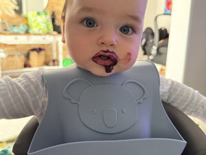 Baby wearing a gray bib with a bear design, sitting in a high chair indoors.