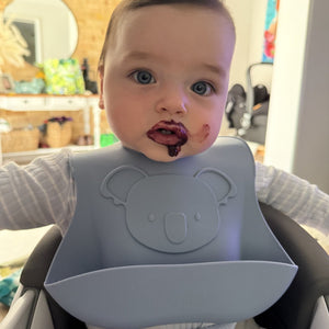 Baby wearing a gray bib with a bear design, sitting in a high chair indoors.