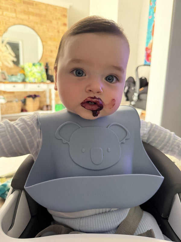 Baby wearing a gray bib with a bear design, sitting in a high chair indoors.