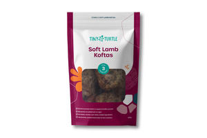 A package of Tiny Turtle Soft Lamb Koitas, stage 3, in a resealable stand-up pouch with a clear window showing the lamb balls inside. The packaging features a white and deep magenta design with orange and teal accents.