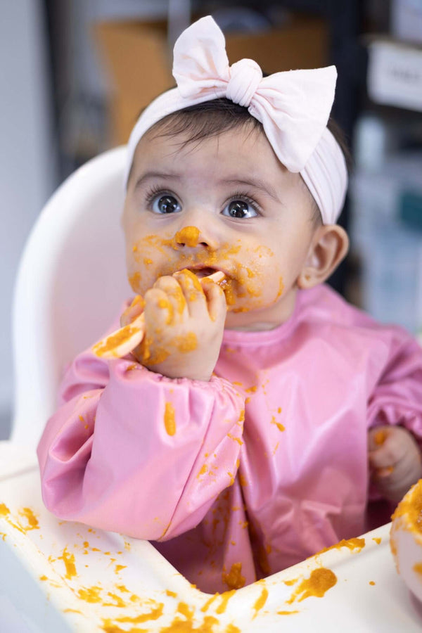 Baby in a high chair with food smudges on face and bib, wearing a pink outfit and headband.