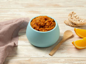 Bowl of Tiny Turtle Prawn & Vegetable Paella baby food with wooden spoon, rice cakes, and orange slices on a wooden surface.