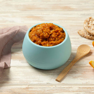 Bowl of Tiny Turtle Prawn & Vegetable Paella baby food with wooden spoon, rice cakes, and orange slices on a wooden surface.