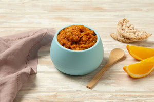 Bowl of Tiny Turtle Prawn & Vegetable Paella baby food with wooden spoon, rice cakes, and orange slices on a wooden surface.